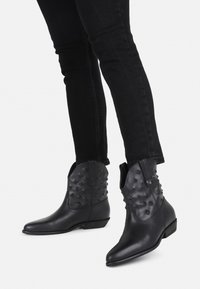Black leather ankle boots featuring studded detailing, pointed toe, and a low stacked heel. Paired with black fitted jeans.