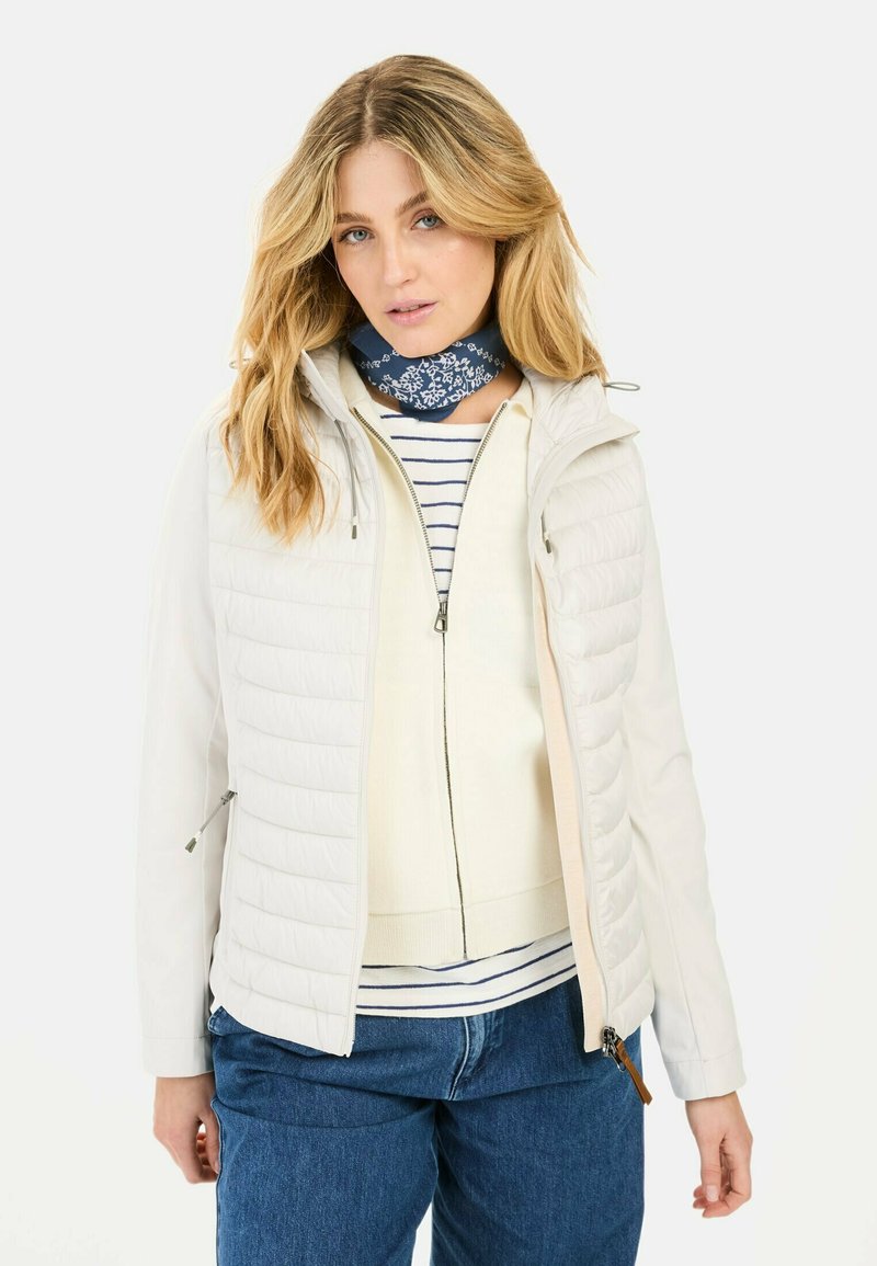 Witte gewatteerde jas met ritssluiting, voorzien van een capuchon en een gestikte textuur. Dragen over een crèmekleurige top en een blauw gestreepte sjaal. Blauwe jeans.