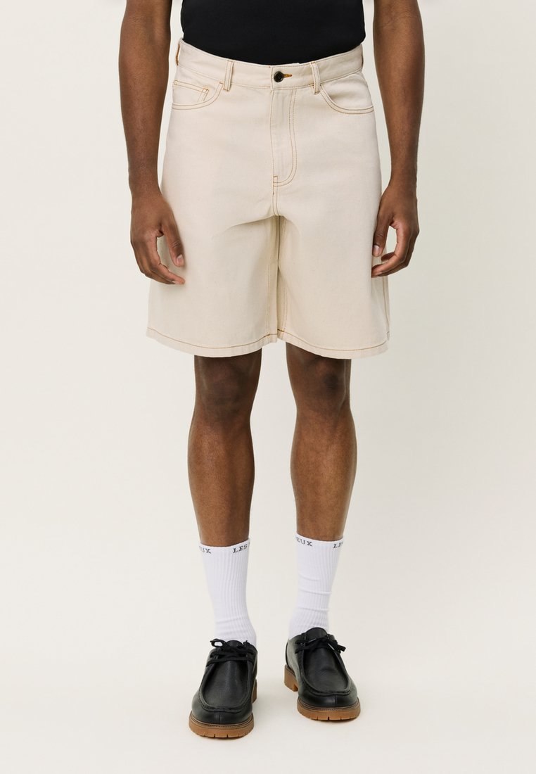 Shorts de mezclilla beige con costuras visibles, diseño clásico de cinco bolsillos y corte recto, combinados con zapatos negros y calcetines blancos.