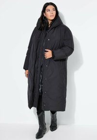 Sort puffer-jakke med høj krave, fuldlængde design, trykknaplukning og lommer, båret over et mønstret indre lag og sorte leggings.