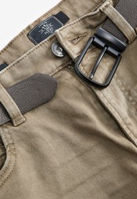Pantaloni di tessuto beige con finitura testurizzata, caratterizzati da una fibbia in metallo nero, una cintura in pelle marrone e un'etichetta cucita all'interno della cintura.