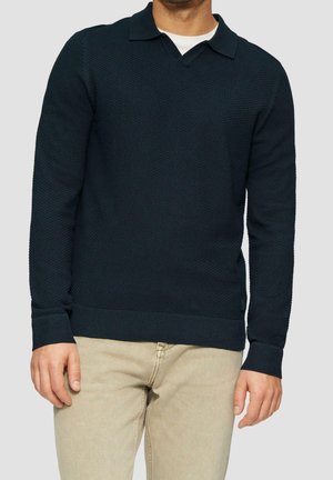 Homme portant un pull polo à manches longues texturé bleu marine par-dessus une chemise blanche avec un pantalon beige, debout devant un fond clair uni.