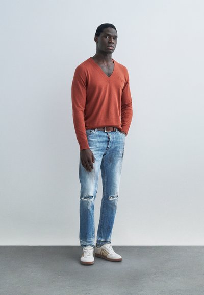 Jeune homme debout, les mains dans les poches, portant un pull orange à col en V, un jean bleu clair déchiré, des baskets blanches, fond gris.