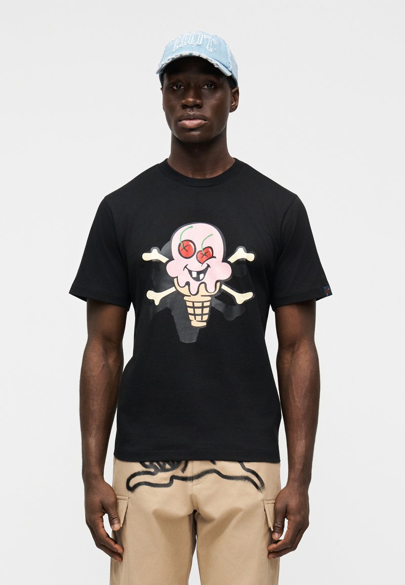 Μαύρο βαμβακερό t-shirt με πολύχρ ωμο σχέδιο παγωτού και οστά κρανίου σε σταυρό. Φορεμένο με μπεζ παντελόνι κάργκο, παρουσιάζοντας μοναδικά σχέδια.