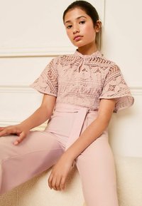 Top de encaje rosa con mangas acampanadas y un cuello alto, combinado con pantalones ajustados de color rosa claro que presentan una cintura amarrada.