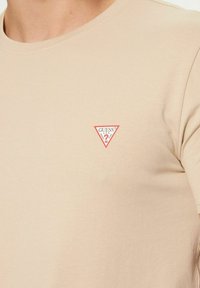 T-shirt in cotone beige con scollo rotondo e piccolo logo triangolare rosso con la scritta "GUESS" sul petto a sinistra. Texture liscia.