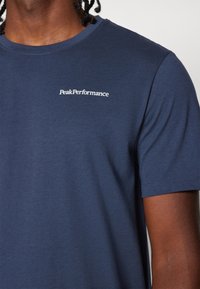 Sötétkék pamut póló kerek nyakkivágással, rövid ujjakkal és fehér "PeakPerformance" logóval a mellkason. Sima textúra.