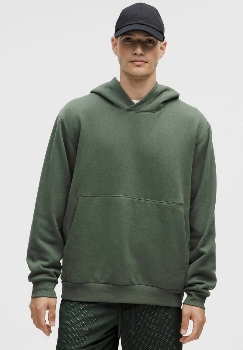 Sudadera con capucha verde oliva hecha de tejido suave, con un bolsillo frontal tipo canguro, puños acanalados y un corte relajado. Usada con una gorra negra.