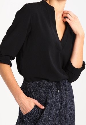Kobieta ubrana w czarną bluzkę z dekoltem w serek i podwiniętymi rękawami oraz ciemne, wzorzyste spodnie, lewa ręka w kieszeni, prawa ręka przy szyi.
