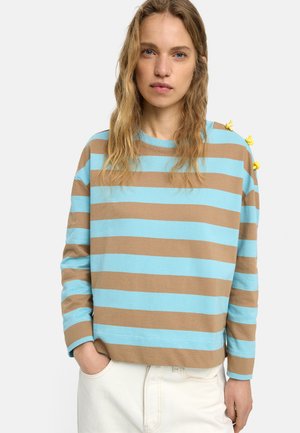 Kobieta o falowanych blond włosach, ubrana w niebiesko-brązową pasiastą koszulę z długim rękawem z żółtymi guzikami na ramieniu oraz jasne spodnie.