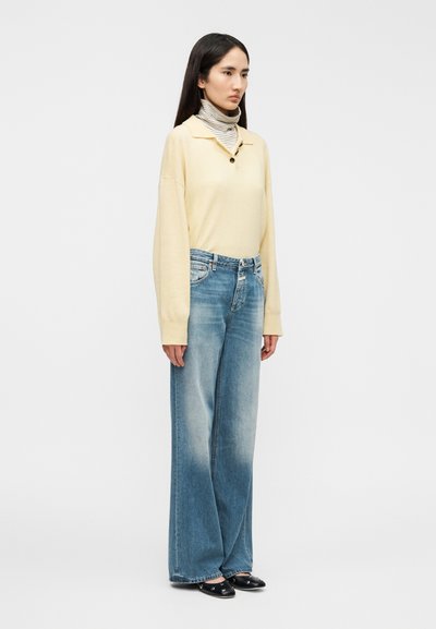 Suéter de manga larga color amarillo pálido, con cuello abotonado, combinado con jeans de pierna ancha en azul claro. Zapatos negros tipo slip-on completan el conjunto.