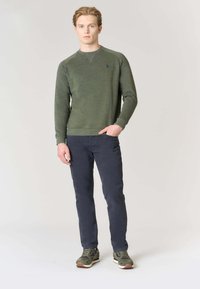 Felpa verde a maniche lunghe con tessuto a trama, caratterizzata da un piccolo logo sul petto, abbinata a pantaloni blu navy e scarpe da ginnastica verdi.