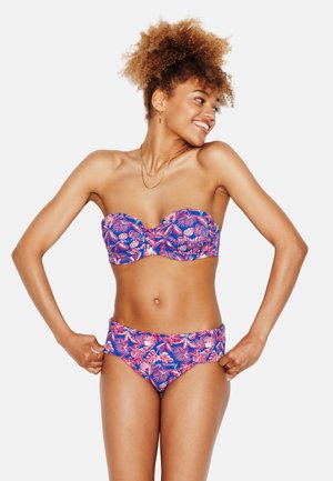 Lachende vrouw die een blauw-roze bloemen strapless bikini verstelt, staand tegen een effen witte achtergrond, kijkend naar rechts.