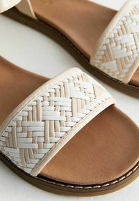 Sandale à glissière tissée blanche avec une large sangle, comportant un motif texturé et une semelle en simili cuir tan avec des détails de couture subtils.