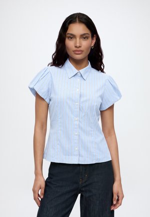 Femme aux longs cheveux foncés portant une chemise à manches courtes rayée bleu et blanc et un jean foncé sur un fond uni.