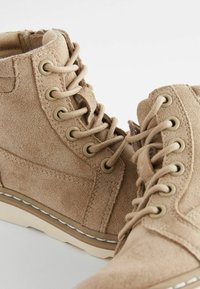 Beige nahka nilkkat bootsit, joissa on metalliset rei'itykset ja yhteensopivat nauhat. Kermanvärinen kumipohja, jossa on valkoiset ommellut yksityiskohdat. Texturoitu pinta, pyöreä kärki.