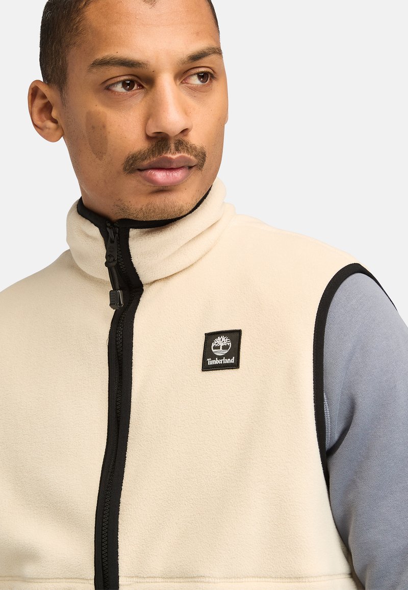 Gilet in pile beige con colletto alto e zip nera, caratterizzato da una patch del logo Timberland nera sul lato sinistro del petto e dettagli in tessuto grigio.