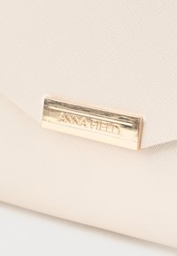 Beige tekstureret clutch-taske med en metalisk guldfarvet logo-plakette. Slank, rektangulær form med et minimalistisk design og uden synligt hardware.