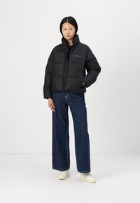 Schwarze gepolsterte Jacke mit hohem Kragen, Reißverschluss und zwei Vordertaschen, kombiniert mit weit geschnittenen Jeans und weißen Sneakers.
