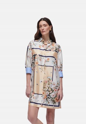 Donna che indossa un vestito floreale a bottoni fino al ginocchio con pannelli beige, bianchi e blu, e polsini a righe blu sulle maniche risvoltate.