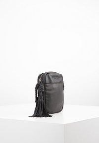 Svart läder crossbody-väska med dragkedjestängning, som har en flätad pom-pom-detalj på sidan och en slät, texturerad yta.