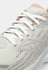 Zapatilla de running gris claro con cordones blancos, parte superior de malla texturizada, sutiles acentos en rosa y un logo en la lengüeta. Presenta un diseño perforado.