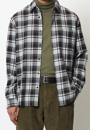 Homme portant un col roulé vert olive, une chemise à carreaux noir et blanc avec boutons, un pantalon en velours côtelé olive et une ceinture noire, debout les mains dans les poches.