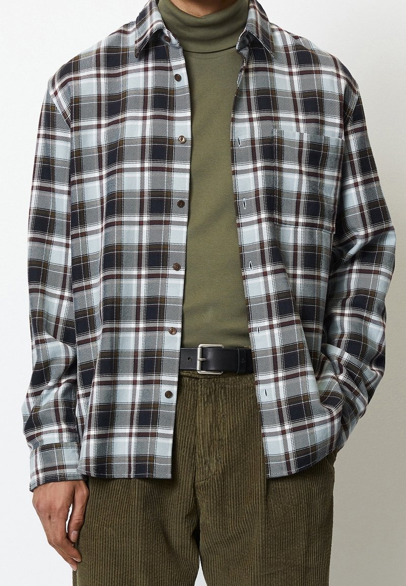 Homme portant un col roulé vert olive, une chemise à carreaux noir et blanc avec boutons, un pantalon en velours côtelé olive et une ceinture noire, debout les mains dans les poches.