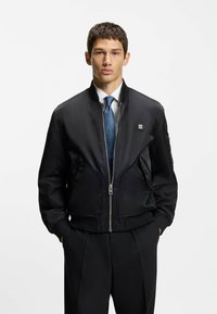 Sort bomberjakke lavet af glat stof, med lynlås foran, ribbet manchetter, to sidelommer og et lille logo på brystet.