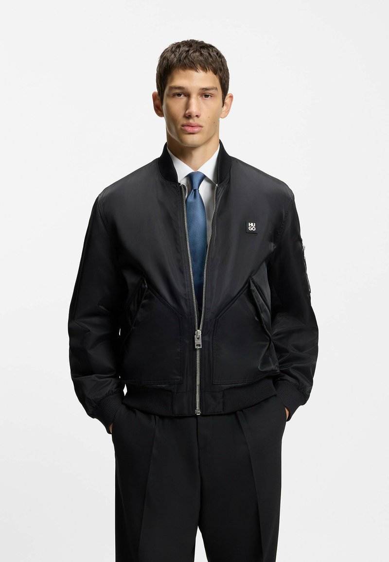 Sort bomberjakke lavet af glat stof, med lynlås foran, ribbet manchetter, to sidelommer og et lille logo på brystet.