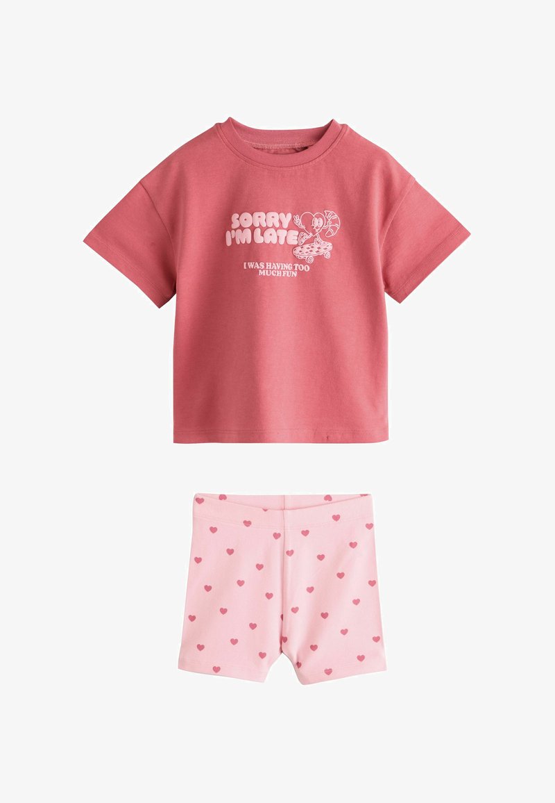 Camiseta rosa de manga corta con dibujo animado blanco y texto combinada con pantalones cortos rosa claro con patrón de pequeños corazones rojos.