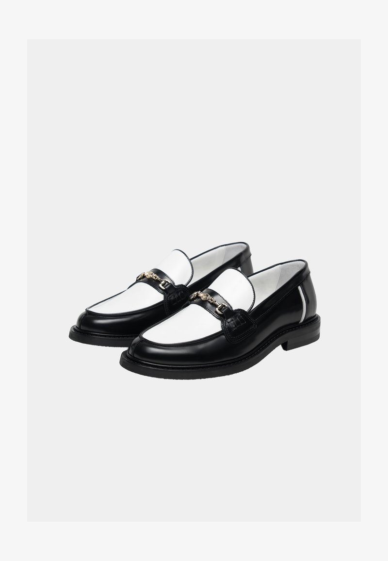 Par sorte og hvide læder loafers med guldkæde på toppen, sat mod en ensartet lys baggrund.