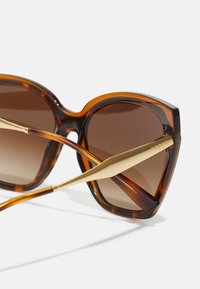 Lunettes de soleil écaille de tortue avec une large monture angulaire, des verres dégradés marron et des accents en métal doré sur les branches.