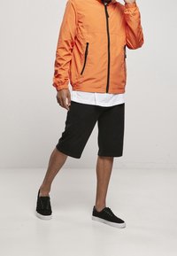 Orange zip-up jakke med sorte detaljer, parret med sorte shorts til knæene og sorte sneakers med hvide såler. Glat stofstruktur.
