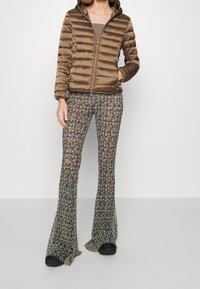 Frau trägt eine braune Daunenjacke mit Kapuze und weit geschnittene, floral gemusterte Hosen, steht vor einem einfarbigen hellen Hintergrund.