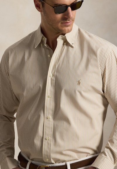 Man draagt een beige-wit verticaal gestreept overhemd met knopen, met geborduurd logo op de borst en donkere zonnebril.