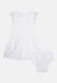 Vit bomullsklänning med volangärmar, broderad detaljer och en rynkad kjol, åtföljd av matchande vita byxor. Dragkedja bak.