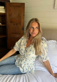 Jeune femme aux longs cheveux blonds portant un chemisier blanc à imprimé floral bleu et un jean bleu, assise sur un lit dans une pièce en bois.
