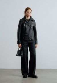 Schwarze Leder-Bikerjacke mit asymmetrischen Reißverschlüssen, tailliert mit Gürtel, kombiniert mit lockeren dunklen Jeanshosen und einer kleinen schwarzen Handtasche.