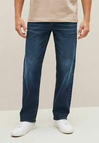 Jeans de mezclilla azul oscuro con un corte recto, que presentan una sutil textura desgastada, detalles de costura y una cinturilla estándar. Combinados con zapatillas blancas.
