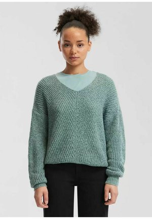 Młoda kobieta o kręconych włosach, ubrana w teksturowany jasnoniebieski sweter z dekoltem w serek na jaśniejszą niebieską bluzkę, stojąca na jednolitym tle.