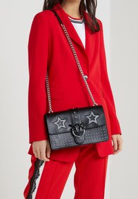 Borsa nera con accenti a stella, borchie e tracolla a catena. Presenta una chiusura a flap con hardware decorativo e finitura testurizzata.