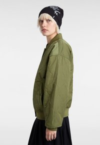 Olive green quilted bomber jacket with a ribbed collar, side pockets, and a smooth texture, paired with a black knit beanie featuring white design.