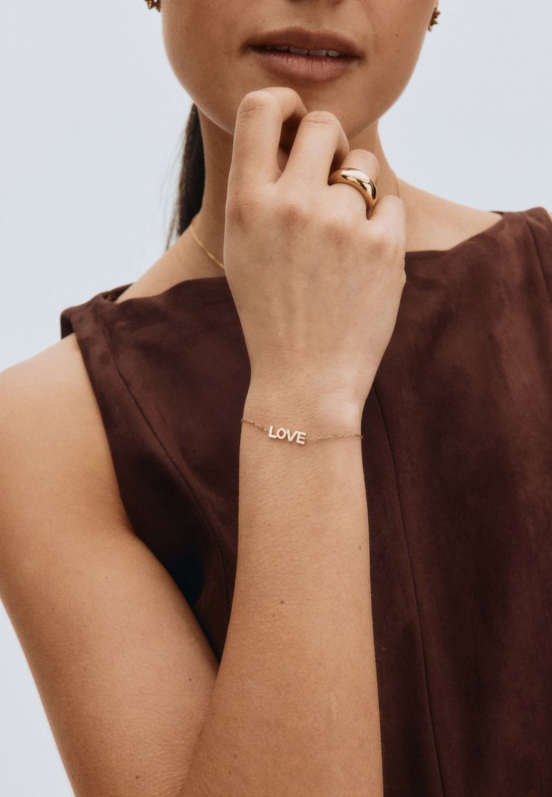 Vrouw met een donker mouwloos topje, die met haar linkerhand haar kin aanraakt, waarbij ze een gouden armband met het woord "LOVE" en een dikke gouden ring draagt.