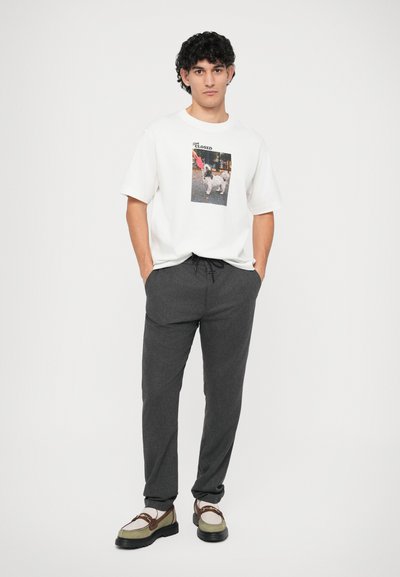 T-shirt branca de manga curta com impressão gráfica e decote redondo, combinada com calças de fato cinza-escuras com cordão e sapatos bicolores.