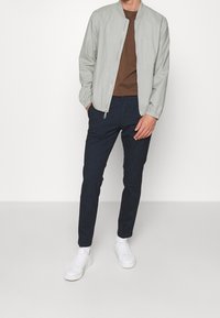 Veste bomber gris clair avec des poignets côtelés, t-shirt marron en dessous, pantalon slim-fit bleu marine et baskets blanches. Design minimaliste et couleurs neutres.
