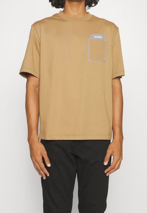 Persoon draagt een beige T-shirt met korte mouwen en een borstzak aan de linkerkant met het label "KORS" en zwarte broek, staand tegen een witte achtergrond.