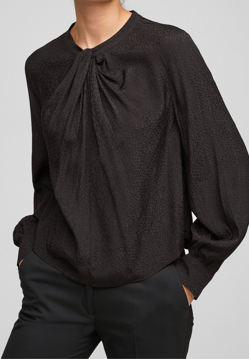 Blusa nera con una trama sottile, caratterizzata da un colletto regolabile, maniche lunghe e un fit rilassato, abbinata a pantaloni neri sartoriali.