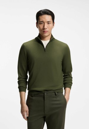 Mann mit schwarzen Haaren, der einen olivgrünen Reißverschluss-Pullover über einem weißen Hemd und passenden olivgrünen Hosen trägt, steht vor weißem Hintergrund.