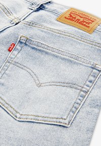 Ανοιχτό μπλε τζιν παντελόνι με τσέπη στο πίσω μέρος διακοσμημένη με καμπύλες ραφές, καφέ δερμάτινο πινακίδα με λογότυπο της μάρκας και μια μικρή κόκκινη ετικέτα.